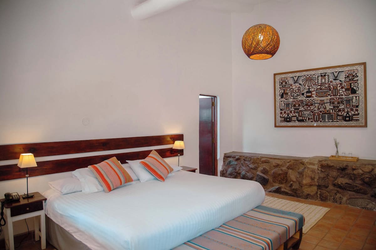 A bedroom with a large bed, two bedside tables, a nightstand, a wall-mounted lamp, a stone accent wall, and a framed artwork.