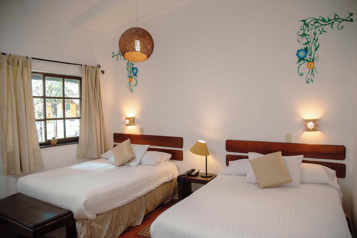 A hotel room with two beds, a window with curtains, a hanging light fixture, and wall-mounted lamps.