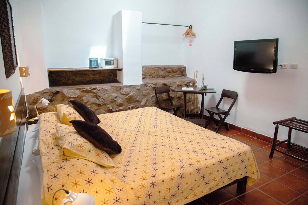 A hotel room with a bed, a small table, a chair, a TV, and a stone wall.