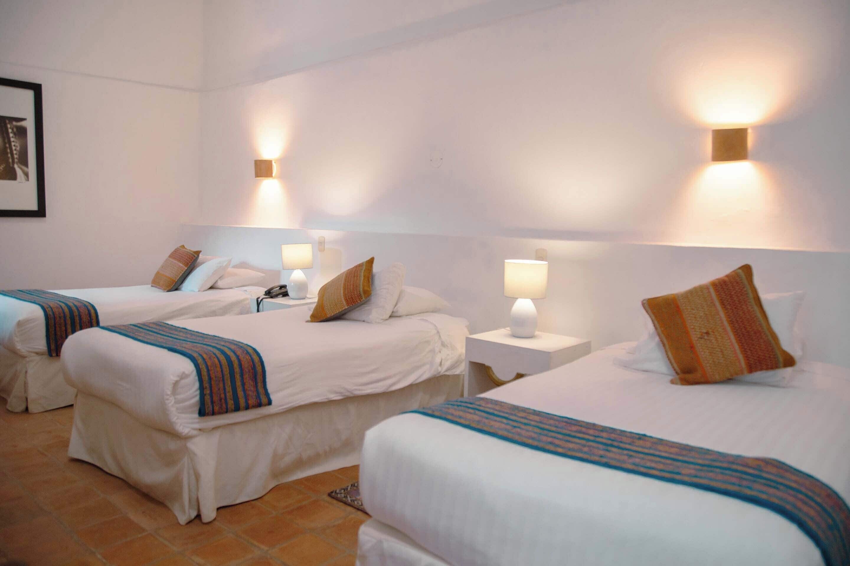 A hotel room with three single beds, each with a bedside table and lamp, and a large bed with a pillow.
