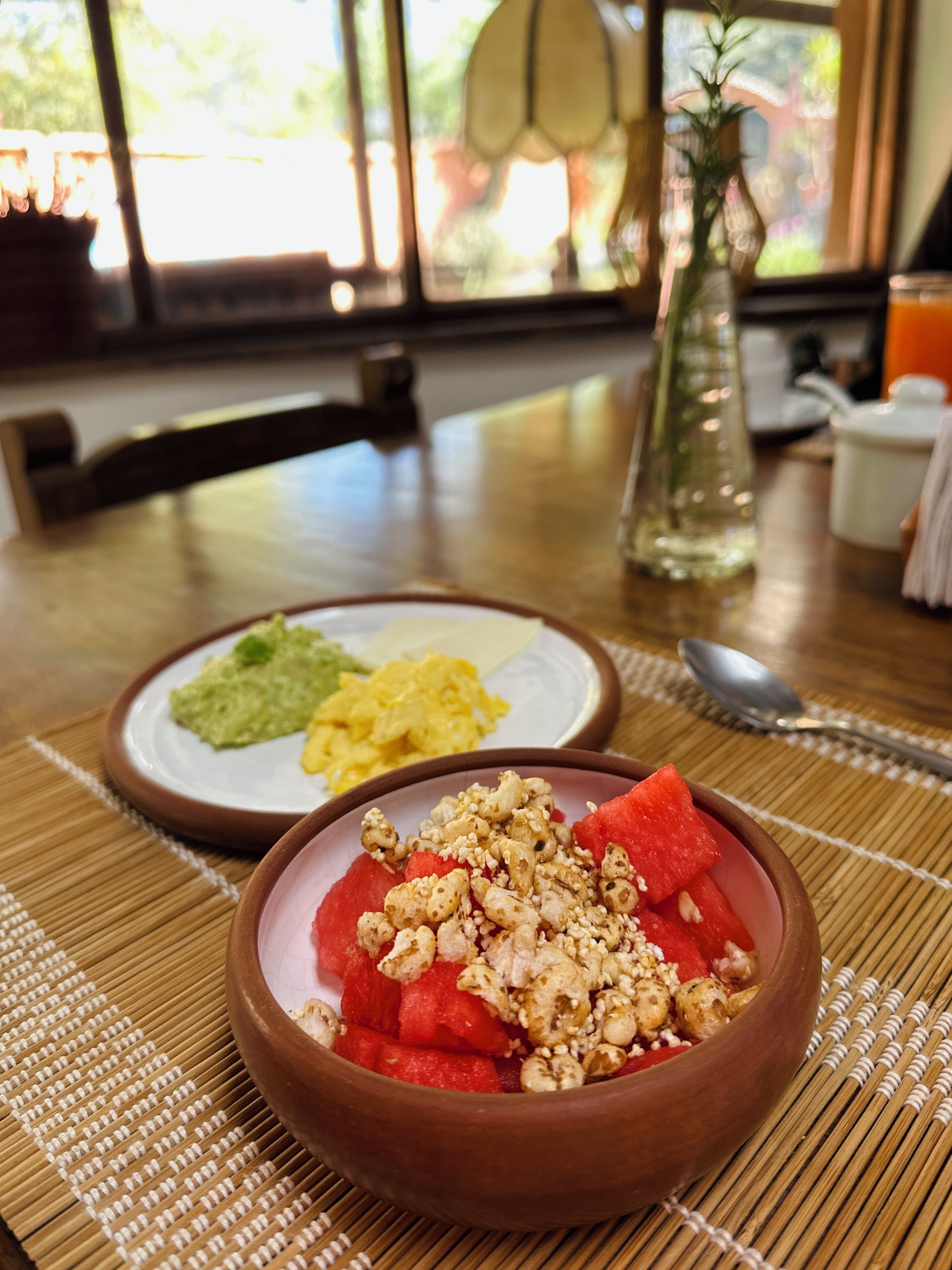 Una mesa de comedor con un tazón de sandía y nueces, un plato de huevos revueltos y aguacate, y un florero con una planta.