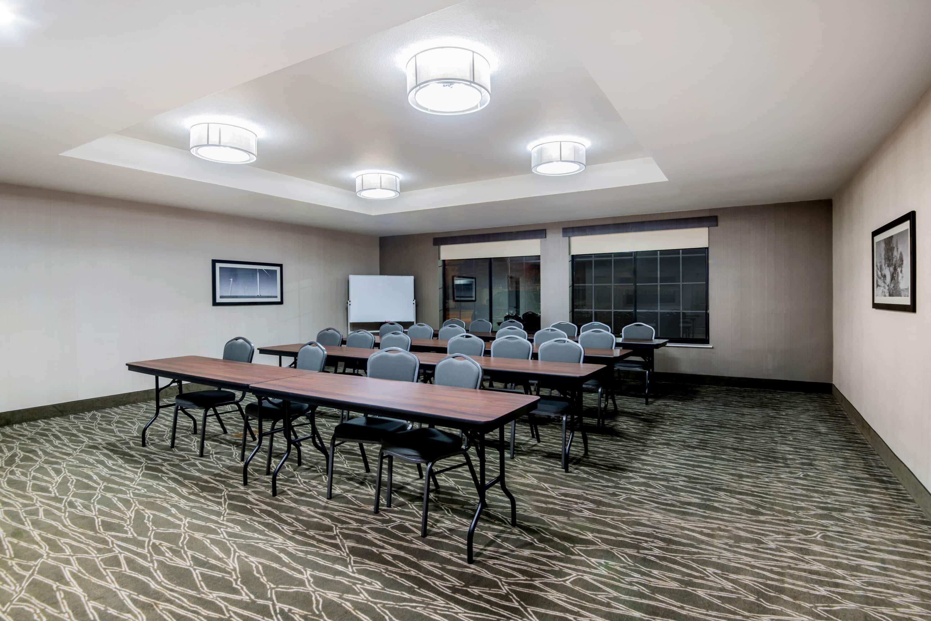 A conference room with a long table, multiple chairs, ceiling lights, a whiteboard, and a window with blinds.