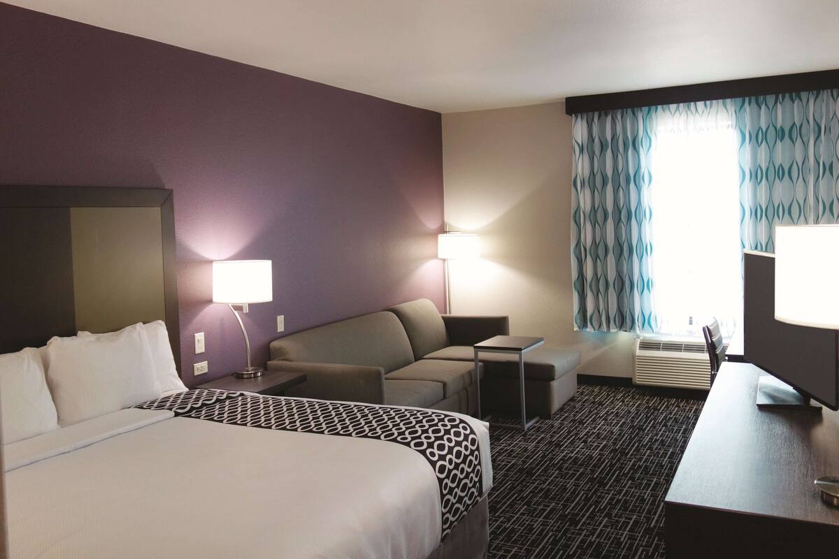 Ett hotellrum med en säng, en soffa, ett skrivbord, en tv och ett fönster med gardiner.