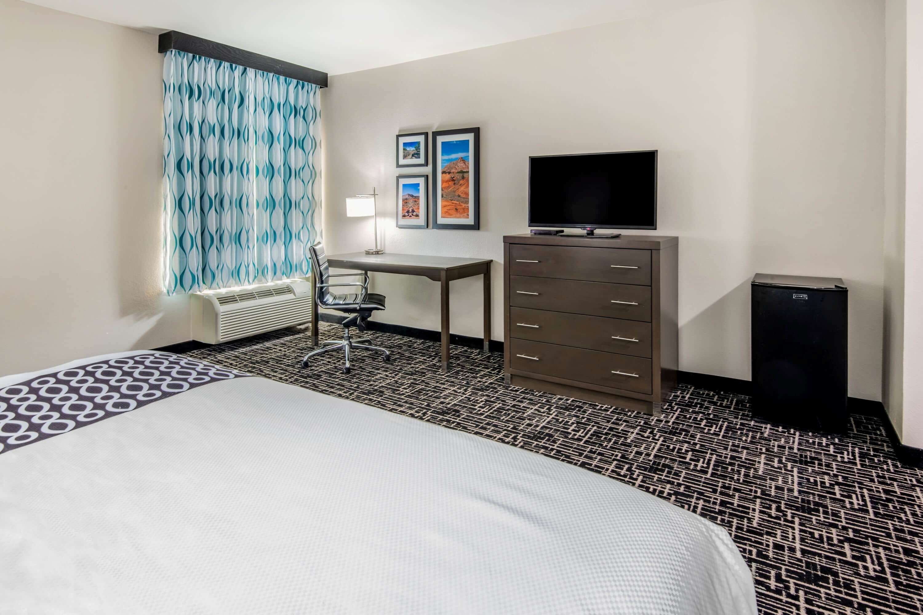 A hotel room with a bed, desk, chair, television, dresser, and a heater.