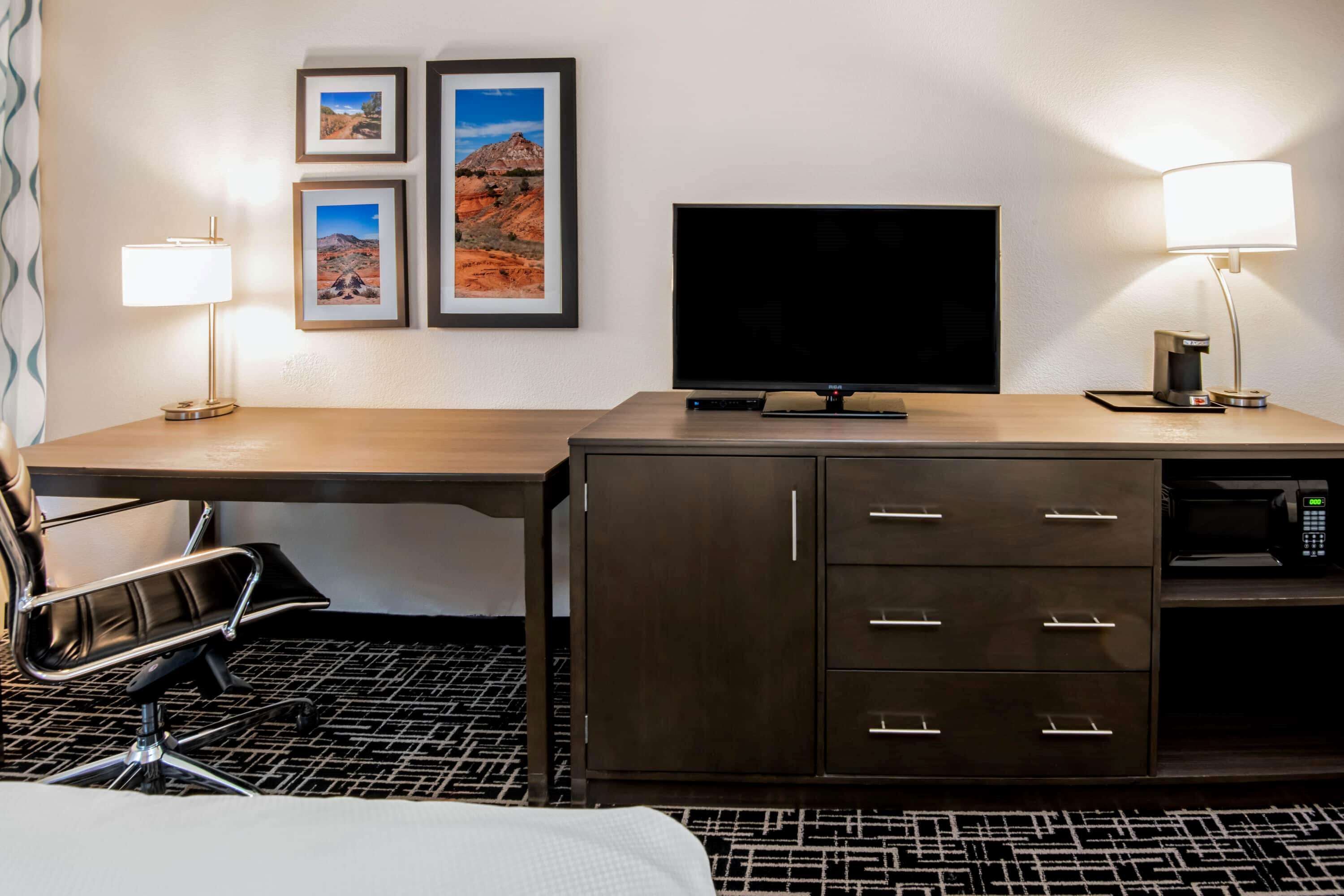 A hotel room with a wooden desk, a black office chair, a flat-screen TV, a minibar, and a bed with a patterned rug.