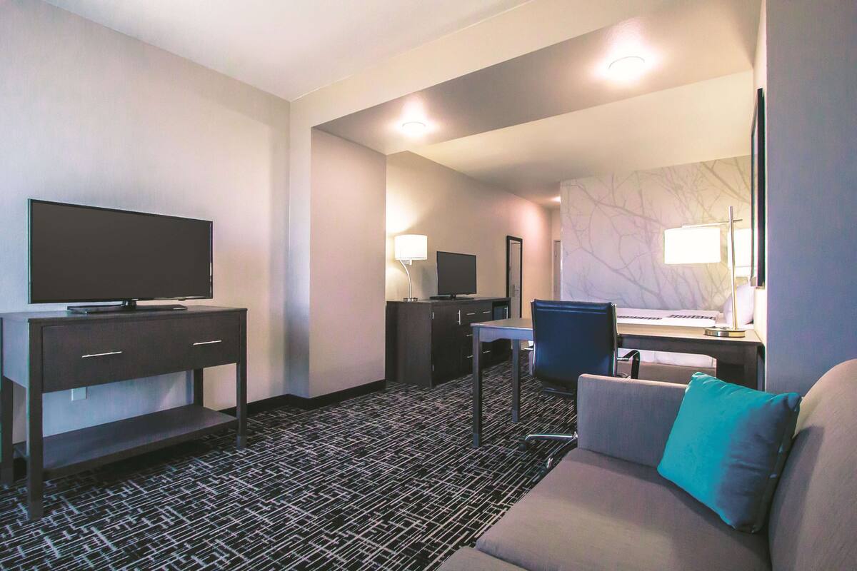 Ett hotellrum med en platt-TV på ett stativ, ett skrivbord med en stol, en soffa med en kudde och en stor spegel på väggen.