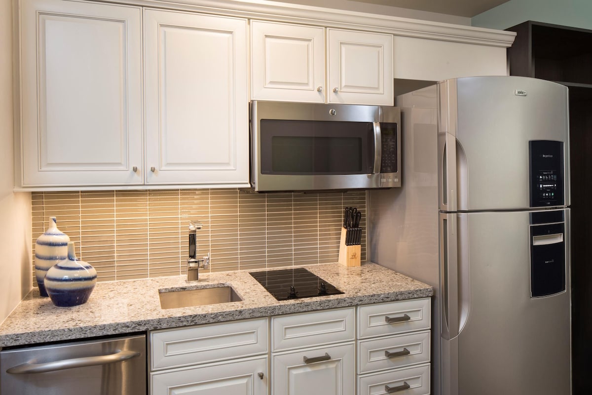 Una cocina moderna con armarios blancos, un refrigerador de acero inoxidable, un microondas integrado, una encimera de granito y un lavavajillas.