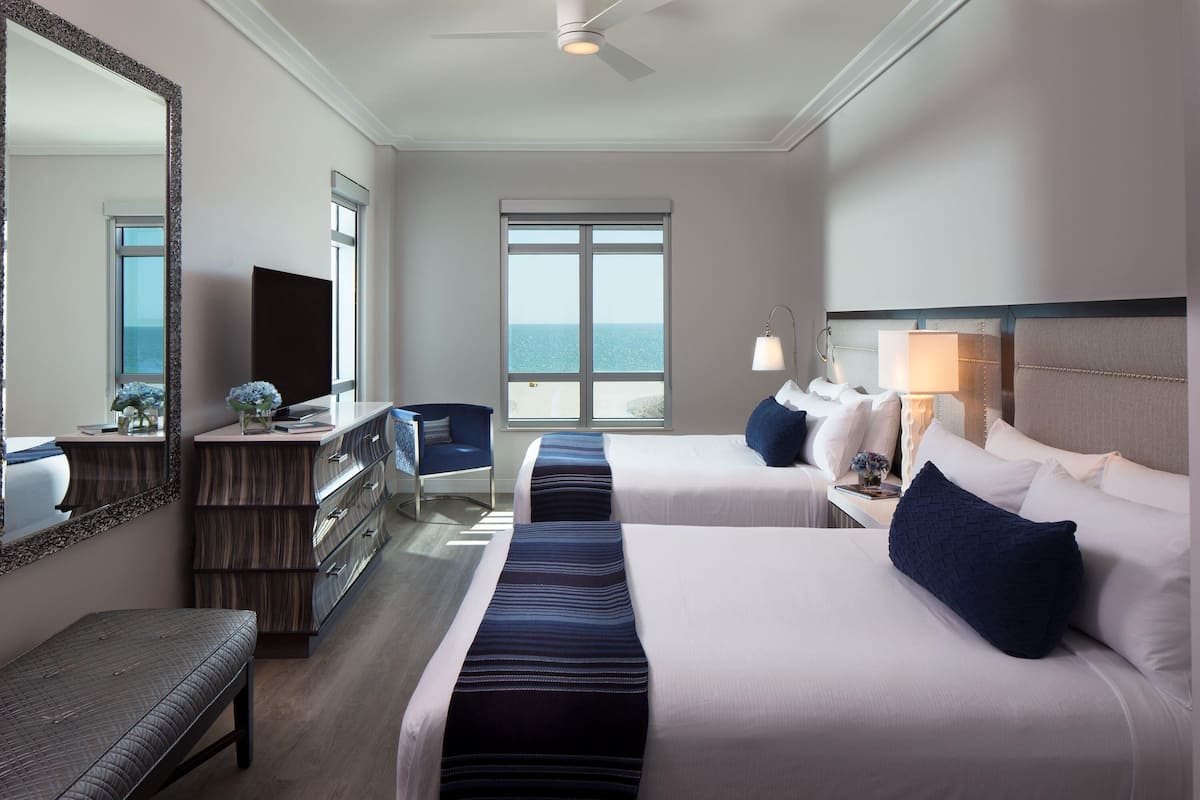 Habitación de hotel con dos camas, un escritorio, una silla, un aparador, un espejo y vista al océano.