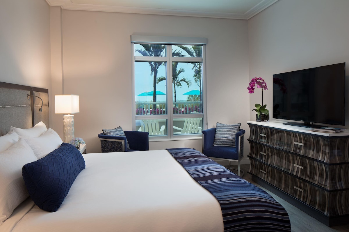 Habitación de hotel con una cama grande, televisión, cómoda y ventana con vista a palmeras.