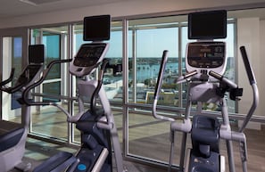 Gimnasio interior con cintas de correr y equipo de ejercicio, amplias ventanas con vistas a un puerto deportivo y edificios.
