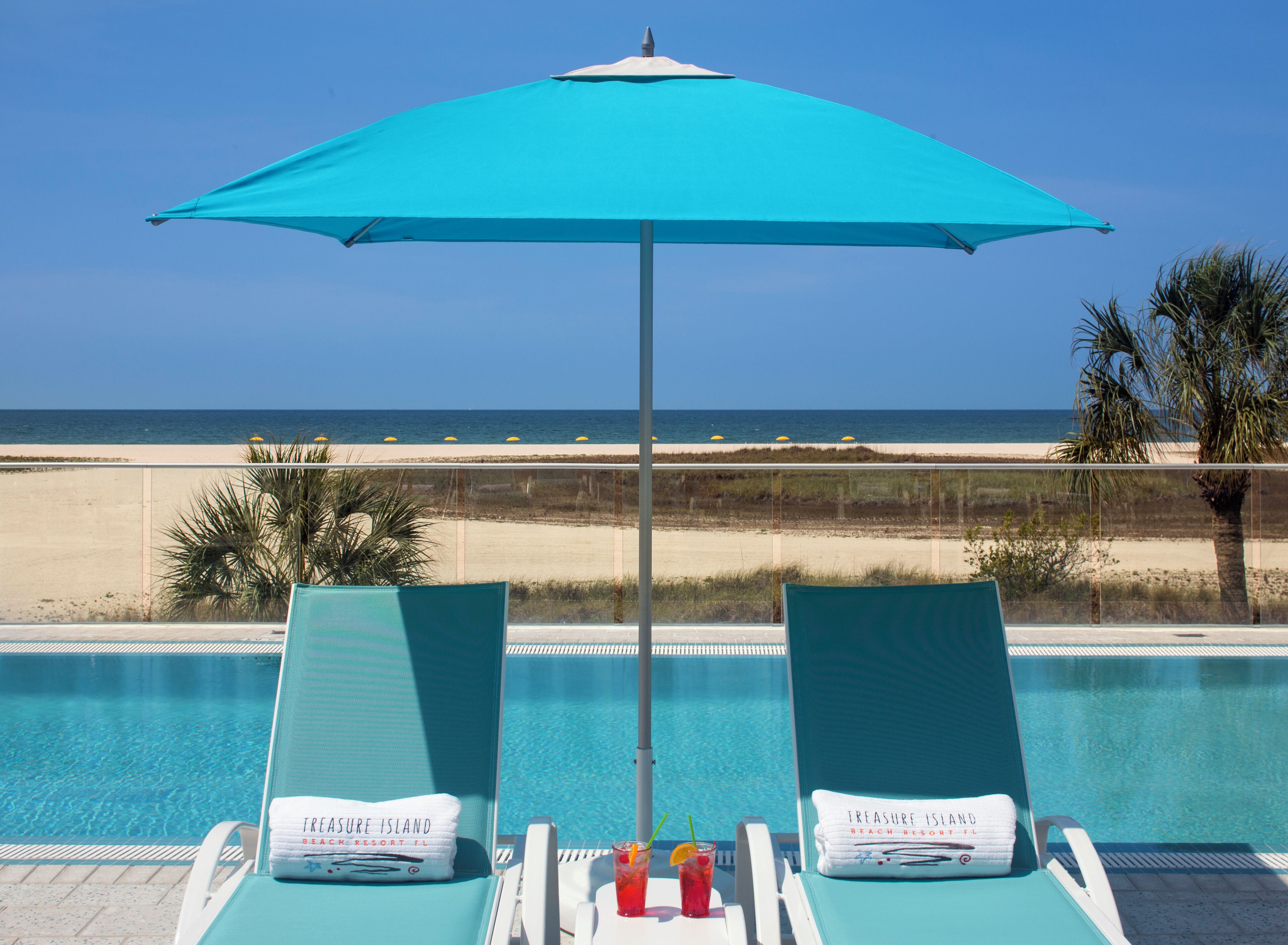 A poolside view with two lounge chairs, a blue umbrella, and a clear view of the beach and ocean.
