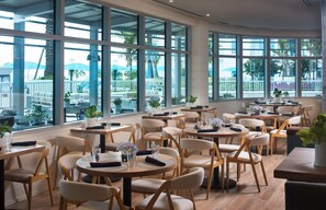 A spacious indoor dining area with multiple round tables and chairs, large windows offering views of the outdoors, and a modern architectural design. - Treasure Island Beach Resort (Treasure Island)