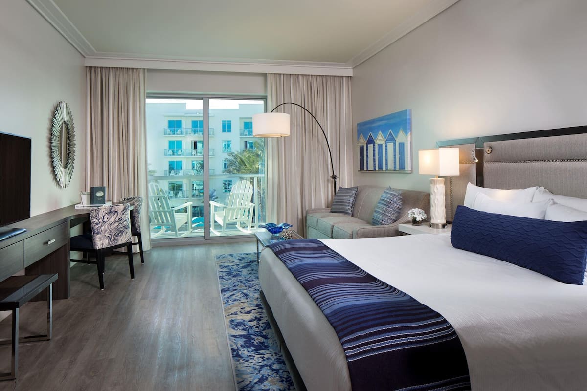 Habitación de hotel con una cama grande, un escritorio con silla, un balcón con vistas a edificios y un cuadro en la pared.