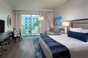 A hotel room with a large bed, a desk with a chair, a balcony with a view of buildings, and a painting on the wall.