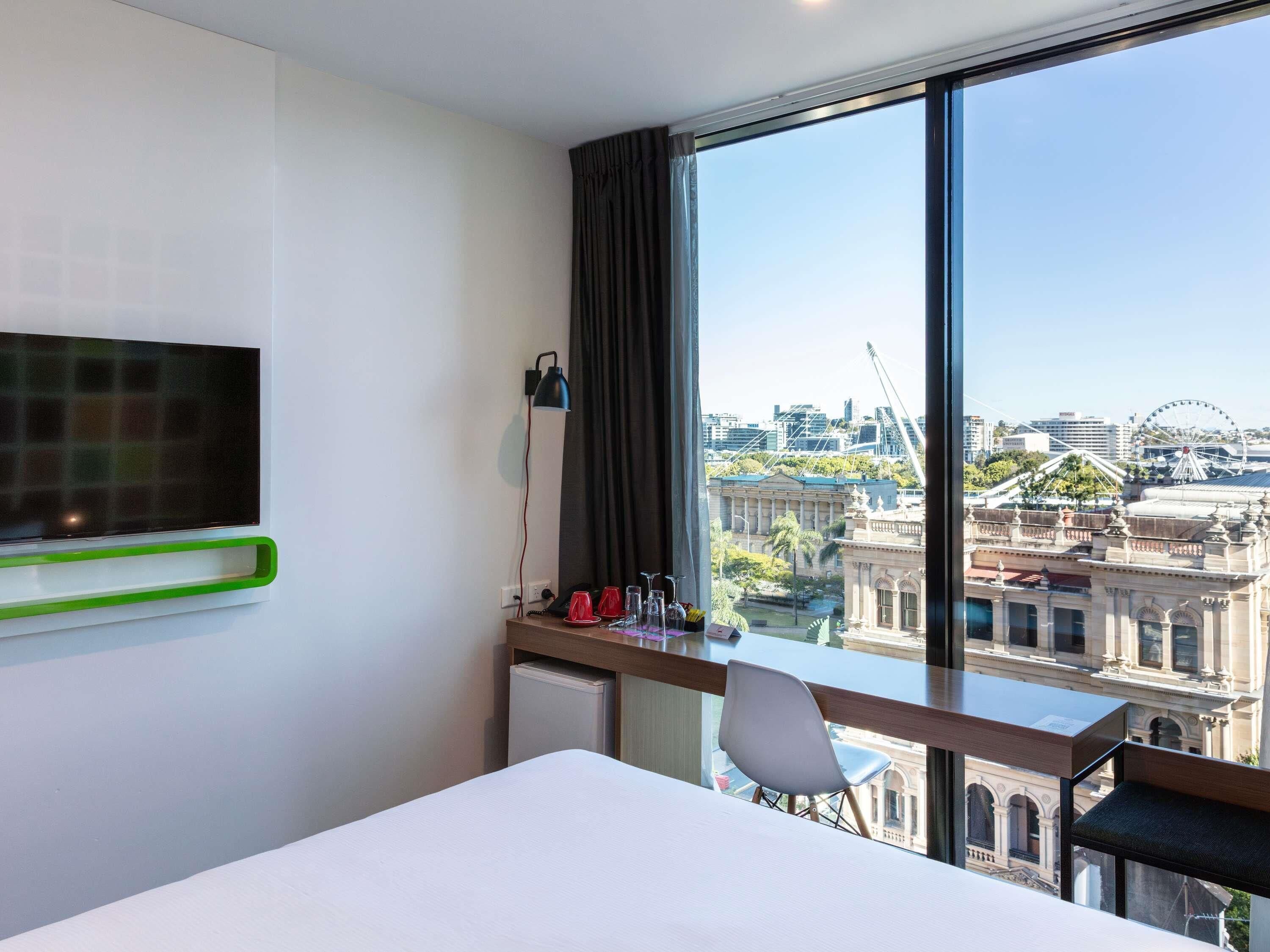 Habitación de hotel con cama, escritorio con silla, televisor montado en la pared, ventana con vistas a la ciudad y lámpara sobre el escritorio.