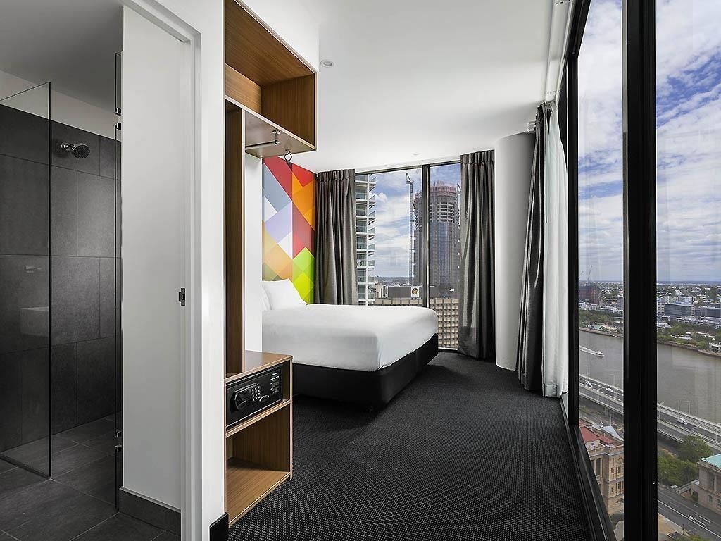 Habitación de hotel moderna con una cama grande, un vestidor amplio, un escritorio pequeño y vistas a la ciudad a través de ventanas panorámicas.