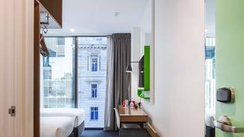 A hotel room with a bed, desk, chair, and a view of buildings through the window.