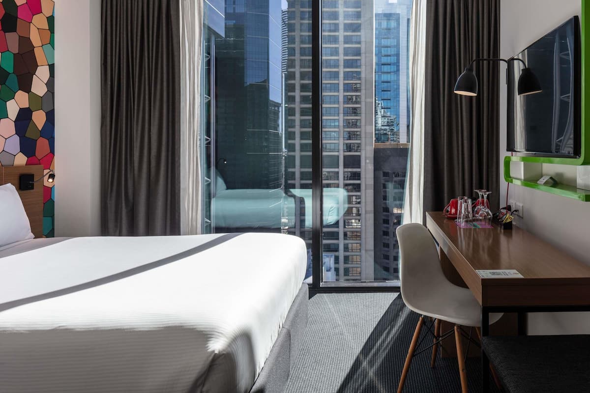 Une chambre d’hôtel avec un lit, un bureau, une chaise, une télévision et une vue sur de hauts immeubles.