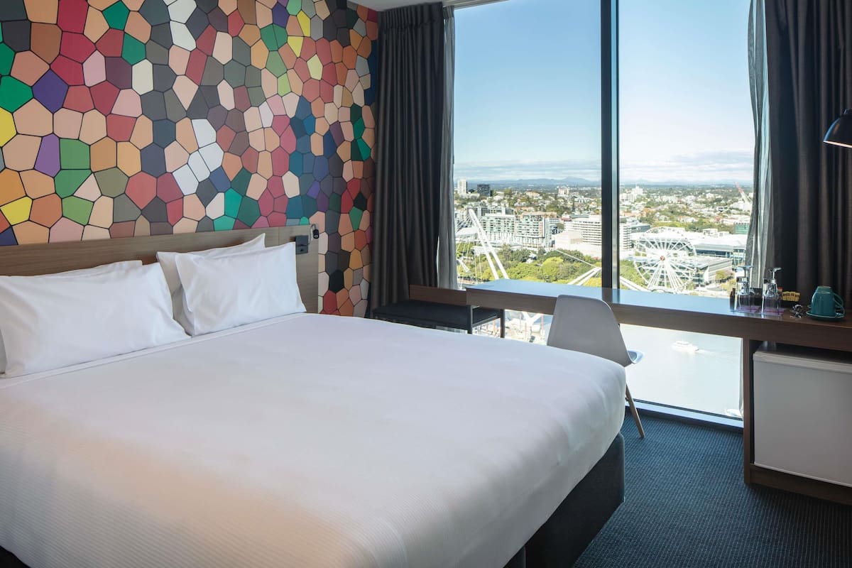 Une chambre d’hôtel dotée d’un grand lit, d’un bureau et offrant une vue sur le paysage urbain grâce à de grandes fenêtres.