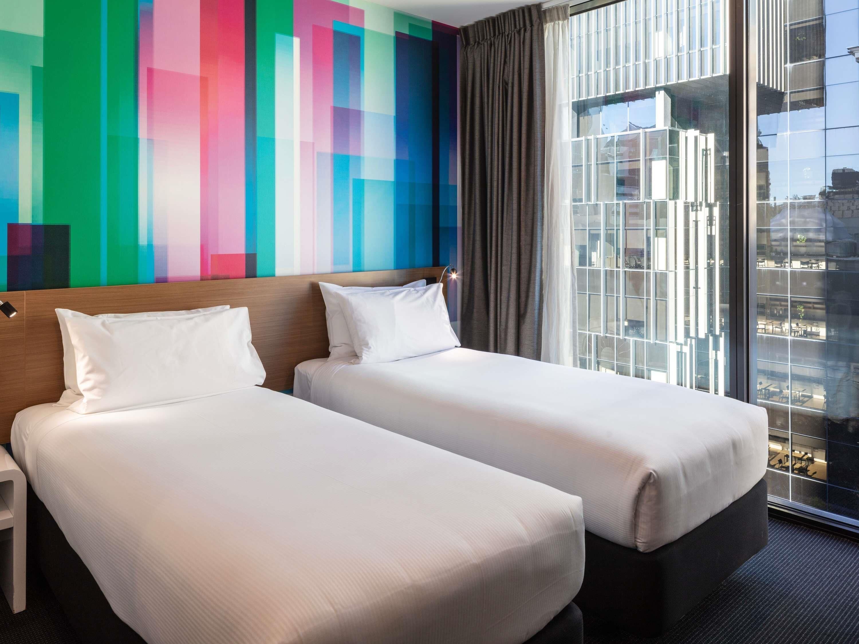 A hotel room with two beds, a colorful wall, a large window with a city view, and a modern design.