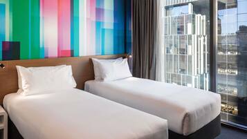 A hotel room with two beds, a colorful wall, a large window with a city view, and a modern design.