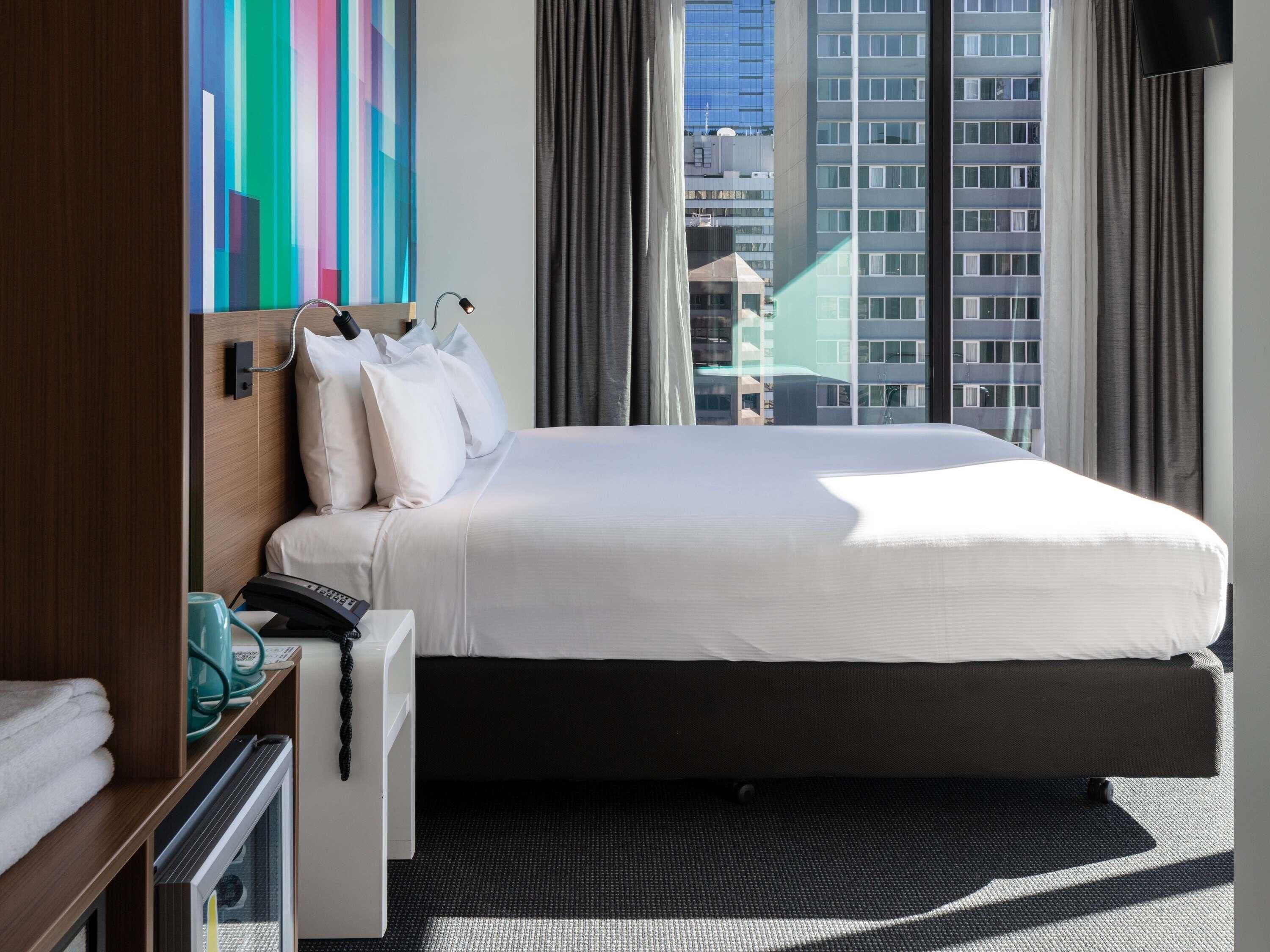 A hotel room with a bed, a desk with a telephone, a television, and a window with a city view.