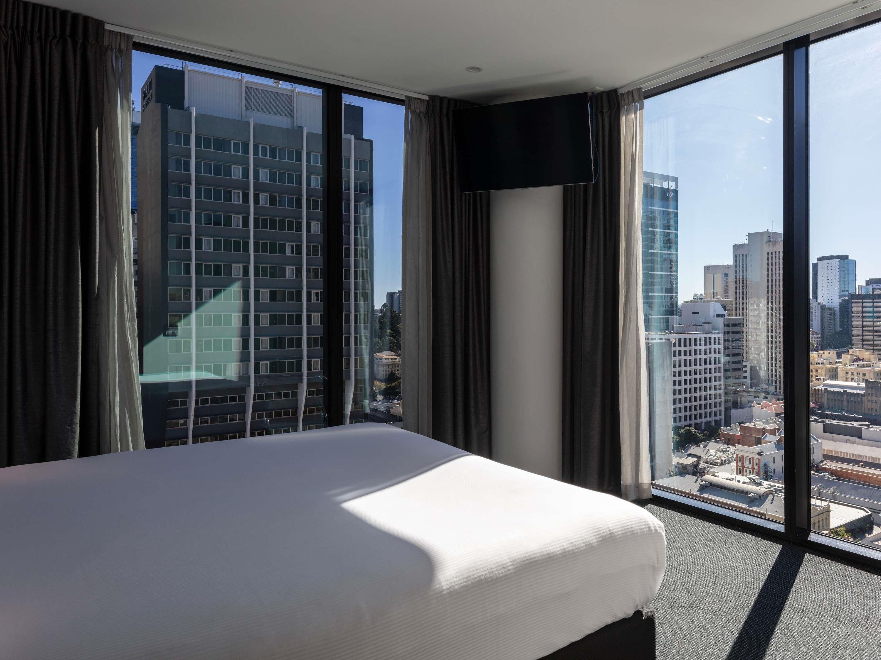 A hotel room with a large bed, a flat-screen TV, and a city view through floor-to-ceiling windows.