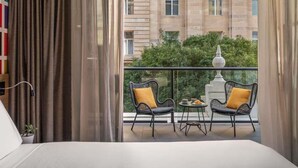 Habitación de hotel con balcón, dos sillas de mimbre con cojines amarillos, una mesita con un plato y una taza, y vistas a un edificio con múltiples ventanas.