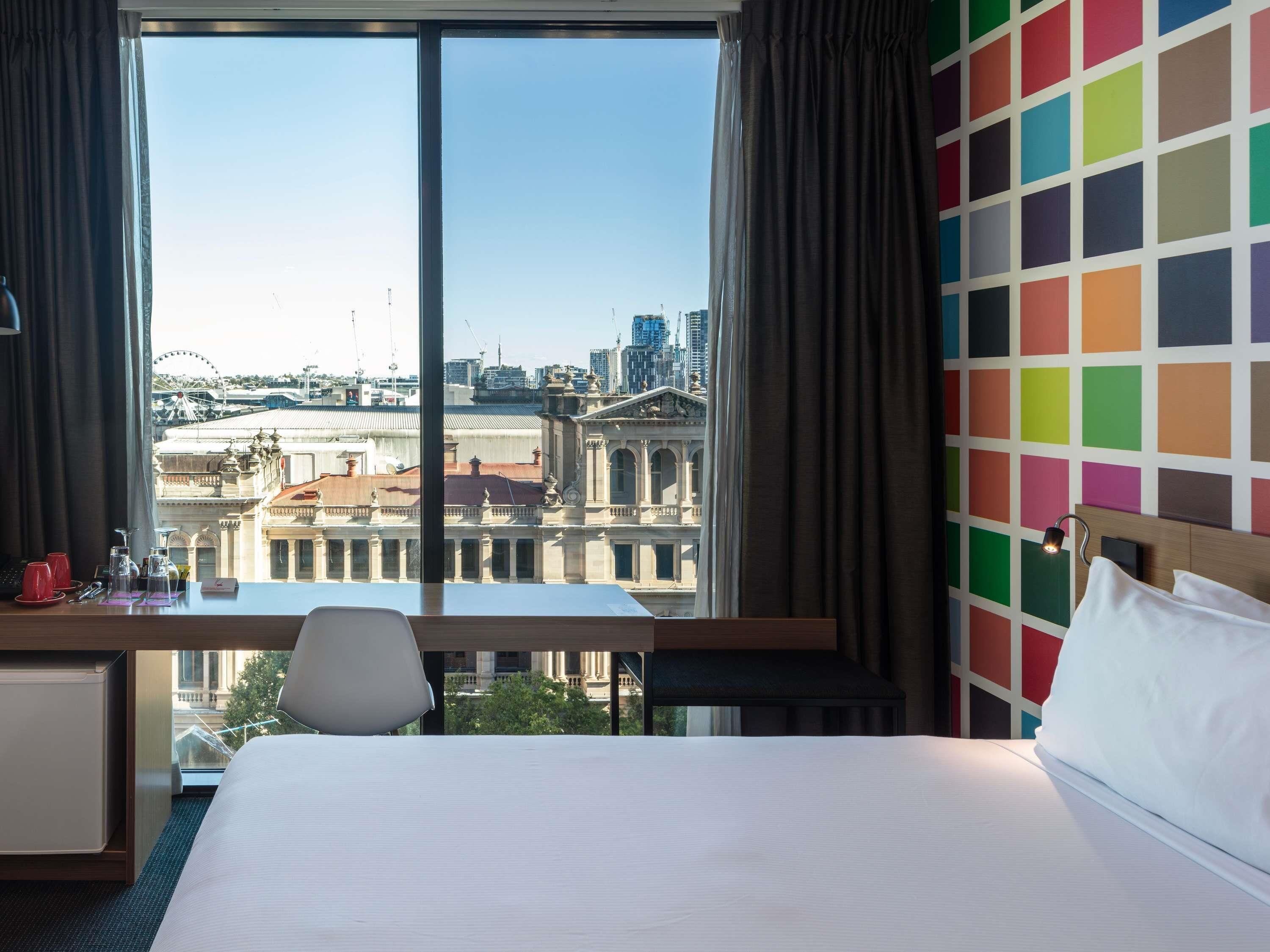 Une chambre d’hôtel avec une grande fenêtre, un bureau, un lit et un mur aux couleurs vives.