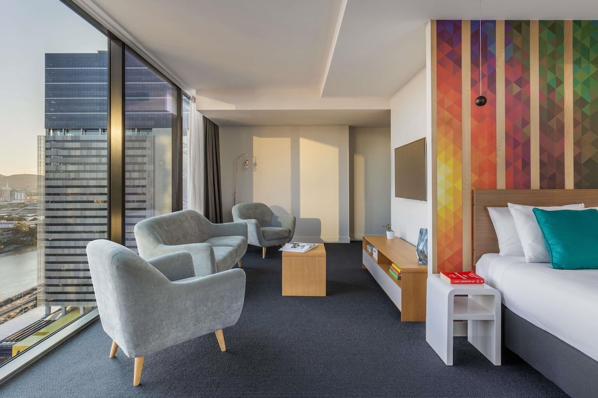 Une chambre d’hôtel avec une grande fenêtre, deux fauteuils, une petite table, un lit et un mur aux couleurs vives.