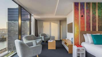 Habitación de hotel con un ventanal grande, dos sillones, una mesita, una cama y una pared colorida.