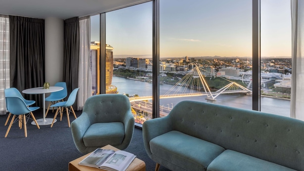 Habitación de hotel con un ventanal grande, un sofá, dos sillas, una mesita y vistas a un paisaje urbano con un puente.