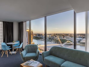 Quarto de hotel com uma janela grande, um sofá, duas poltronas, uma mesa pequena e vista para a paisagem urbana com uma ponte.