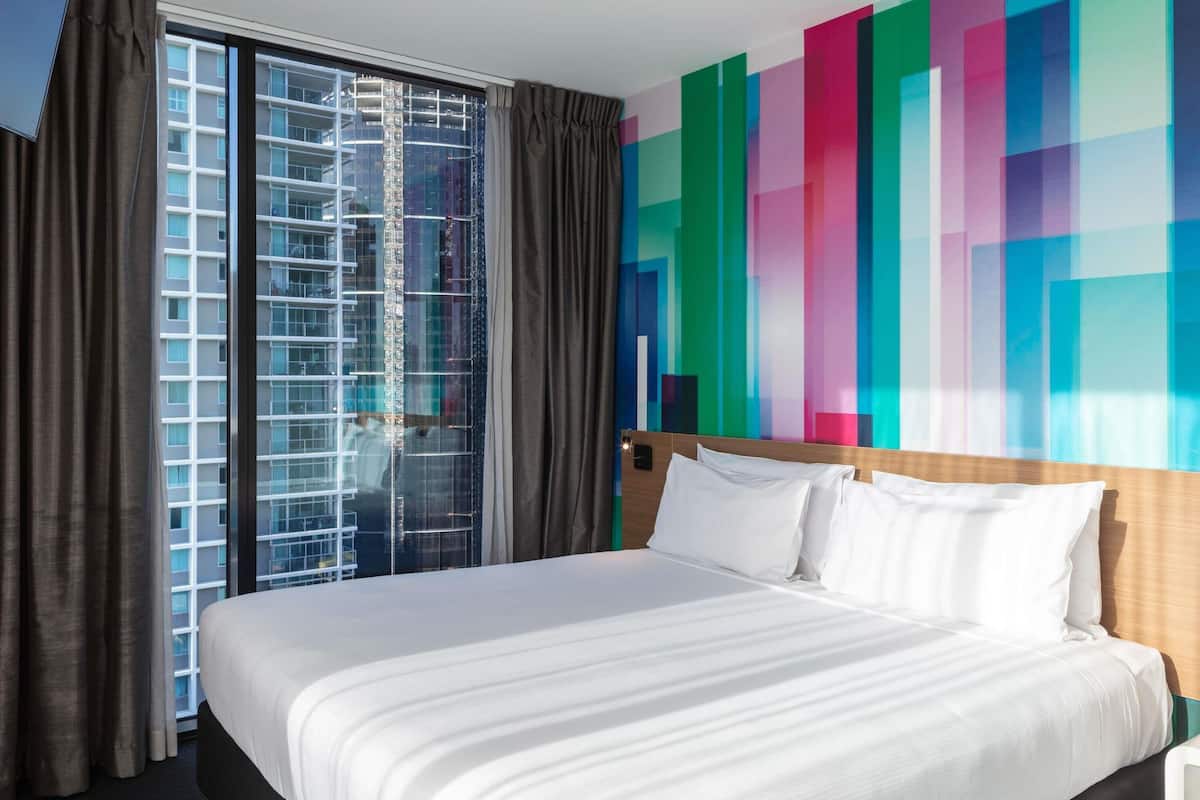 Une chambre d’hôtel avec un grand lit, une vue sur un immeuble par la fenêtre et un mur aux couleurs vives.