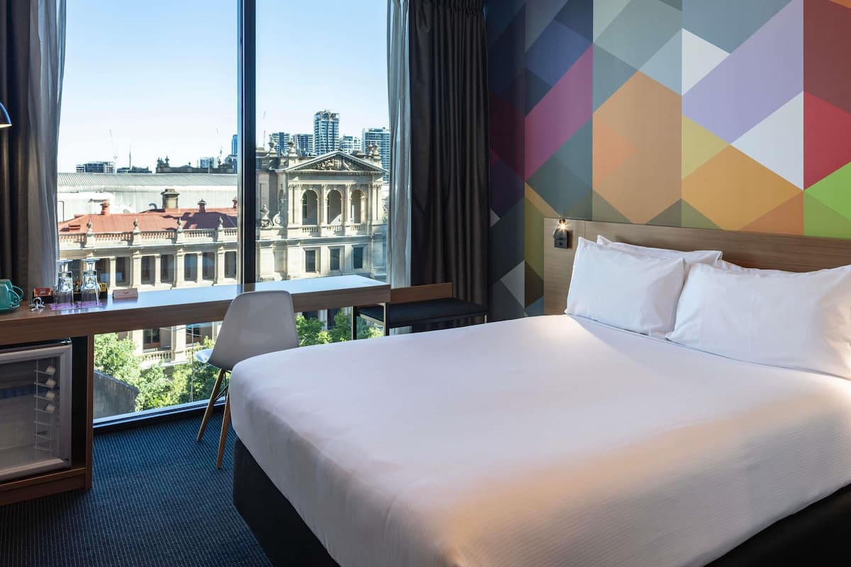 Une chambre d’hôtel avec un grand lit, un bureau avec une chaise et une vue sur le paysage urbain grâce à de grandes fenêtres.