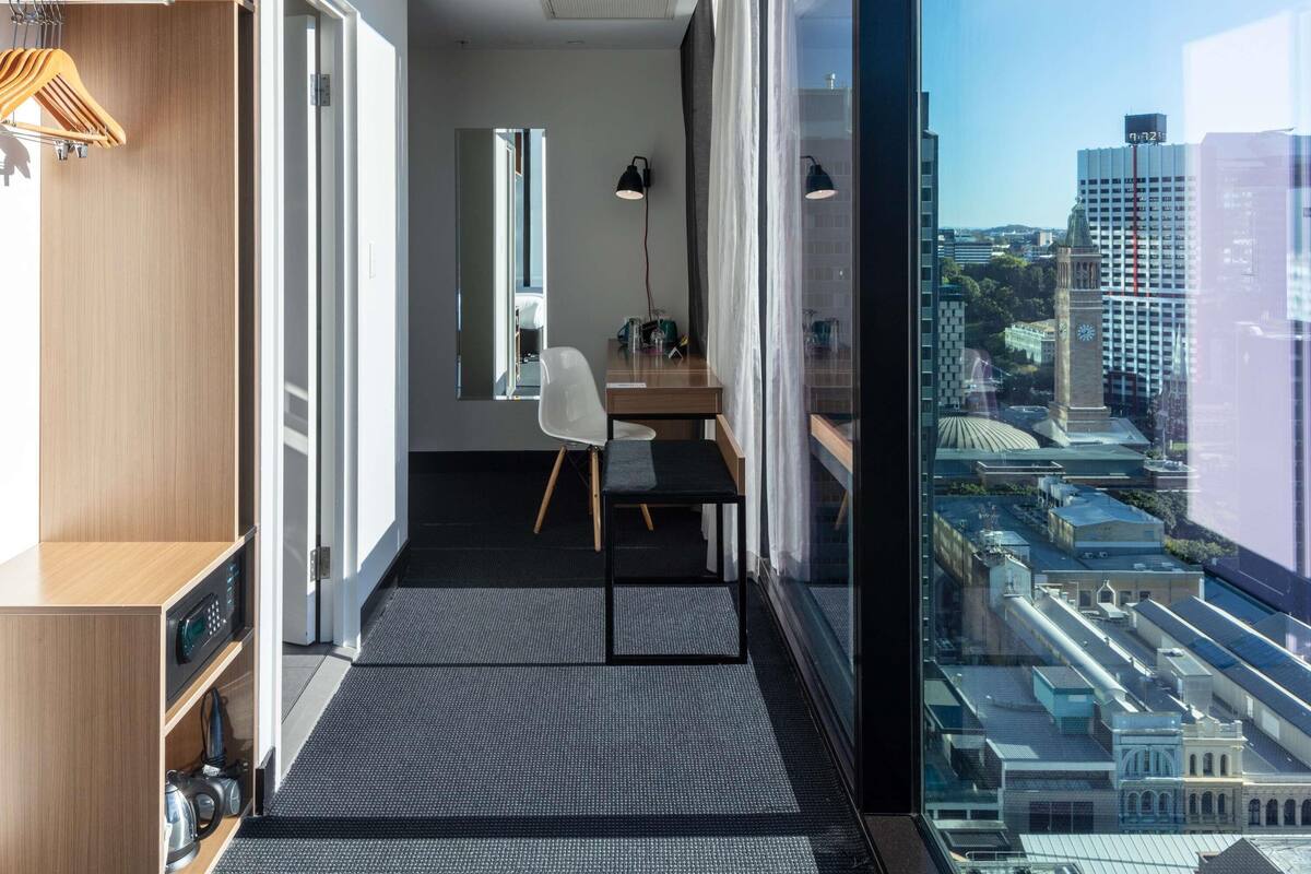 A modern hotel room with a city view, a desk with a chair, a wardrobe, and a large window.