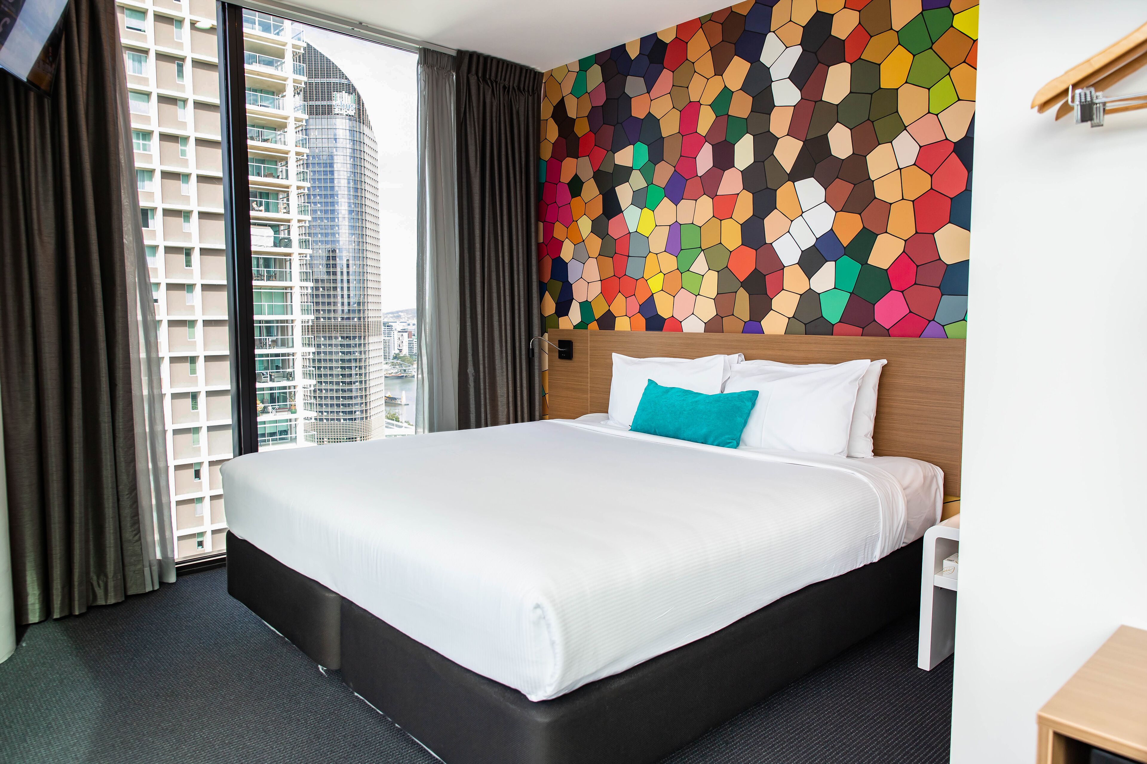 Quarto de hotel com uma cama grande, um painel de mosaicos coloridos e vista para edifícios altos pela janela.