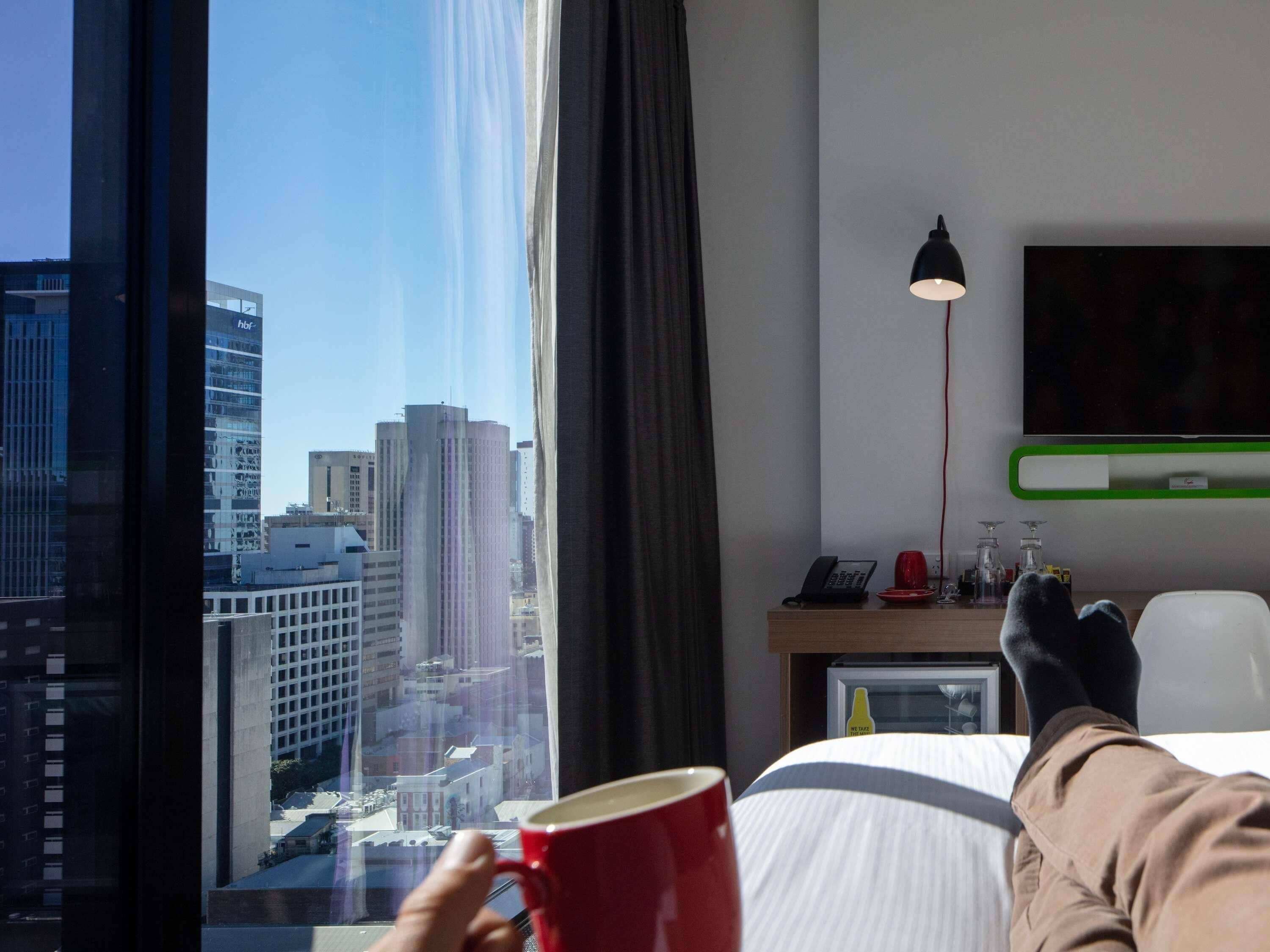A hotel room with a large window, a bed, a desk with a lamp, and a TV on the wall.