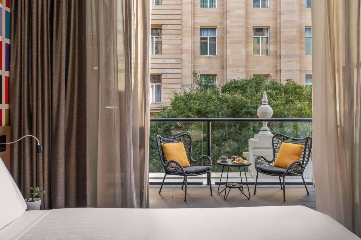 Une chambre d’hôtel avec un balcon, deux fauteuils en osier avec des coussins, une petite table ronde et une vue sur un immeuble et de la verdure.