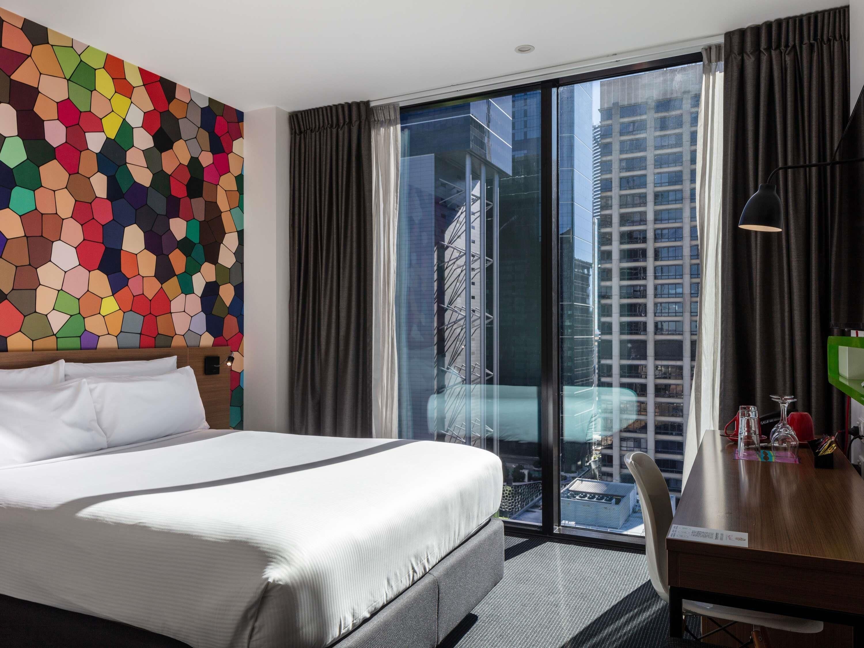 Una habitación de hotel moderna con una cama grande, un diseño hexagonal colorido en la pared, vistas de un paisaje urbano a través de ventanales de piso a techo, y un escritorio con una lámpara y un objeto verde.