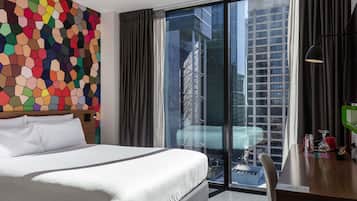 Una habitación de hotel moderna con una cama grande, un diseño hexagonal colorido en la pared, vistas de un paisaje urbano a través de ventanales de piso a techo, y un escritorio con una lámpara y un objeto verde.
