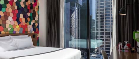 Una habitación de hotel moderna con una cama grande, un diseño hexagonal colorido en la pared, vistas de un paisaje urbano a través de ventanales de piso a techo, y un escritorio con una lámpara y un objeto verde.