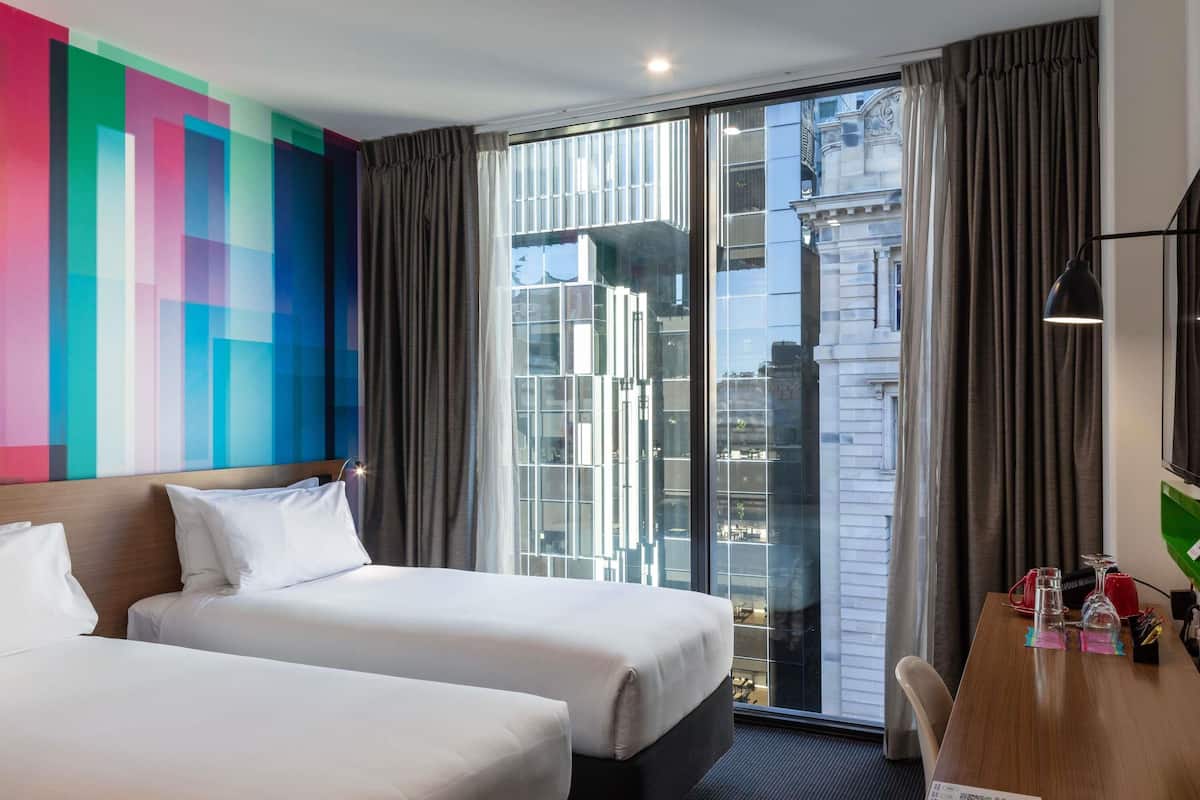 Une chambre d’hôtel avec deux lits, une grande fenêtre donnant sur la ville, un bureau avec une lampe et un mur décoré d’une œuvre abstraite aux couleurs vives.
