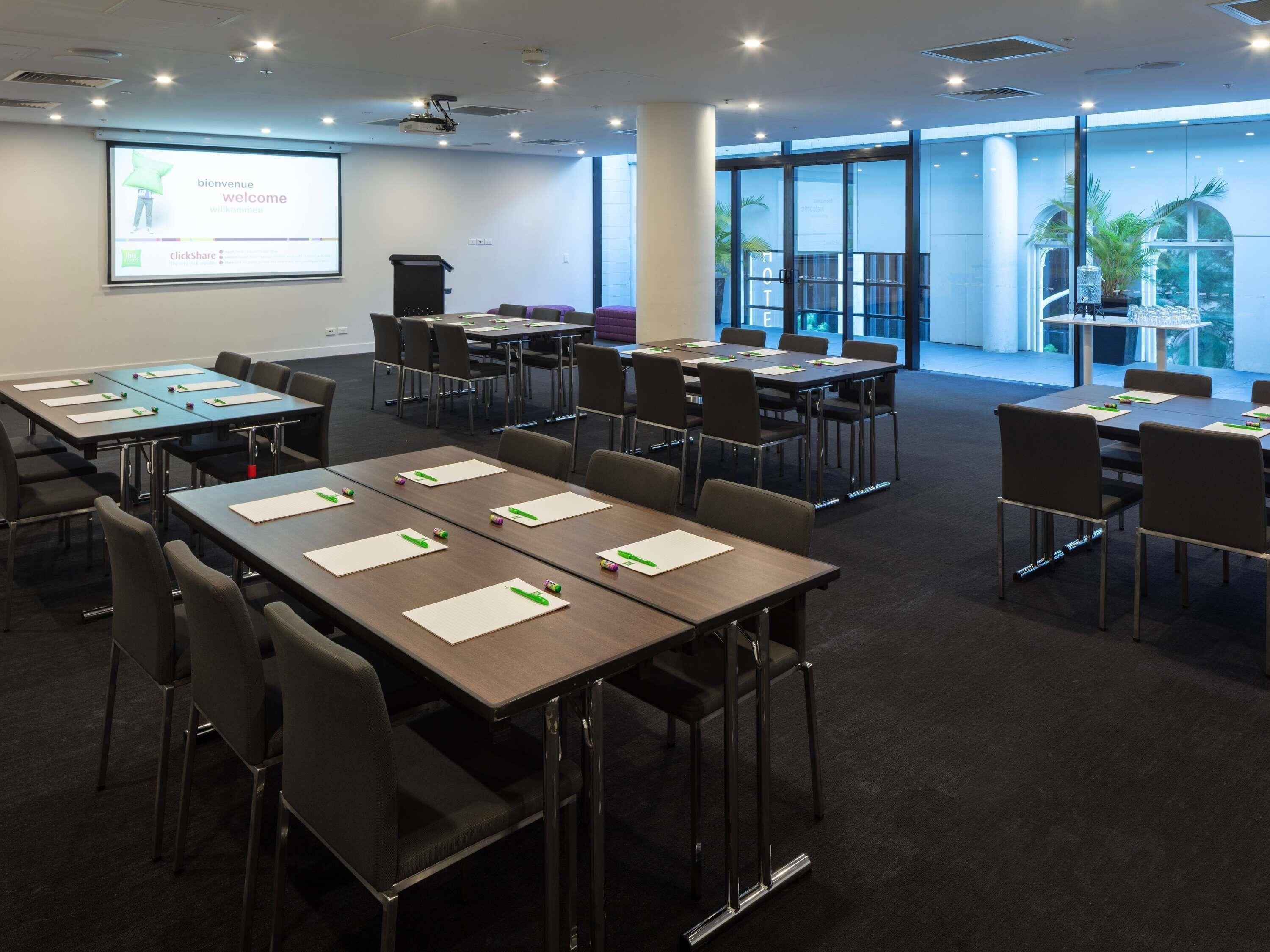 Un salón de conferencias con mesas, sillas, un proyector y una pantalla mostrando 'welcome' en varios idiomas.