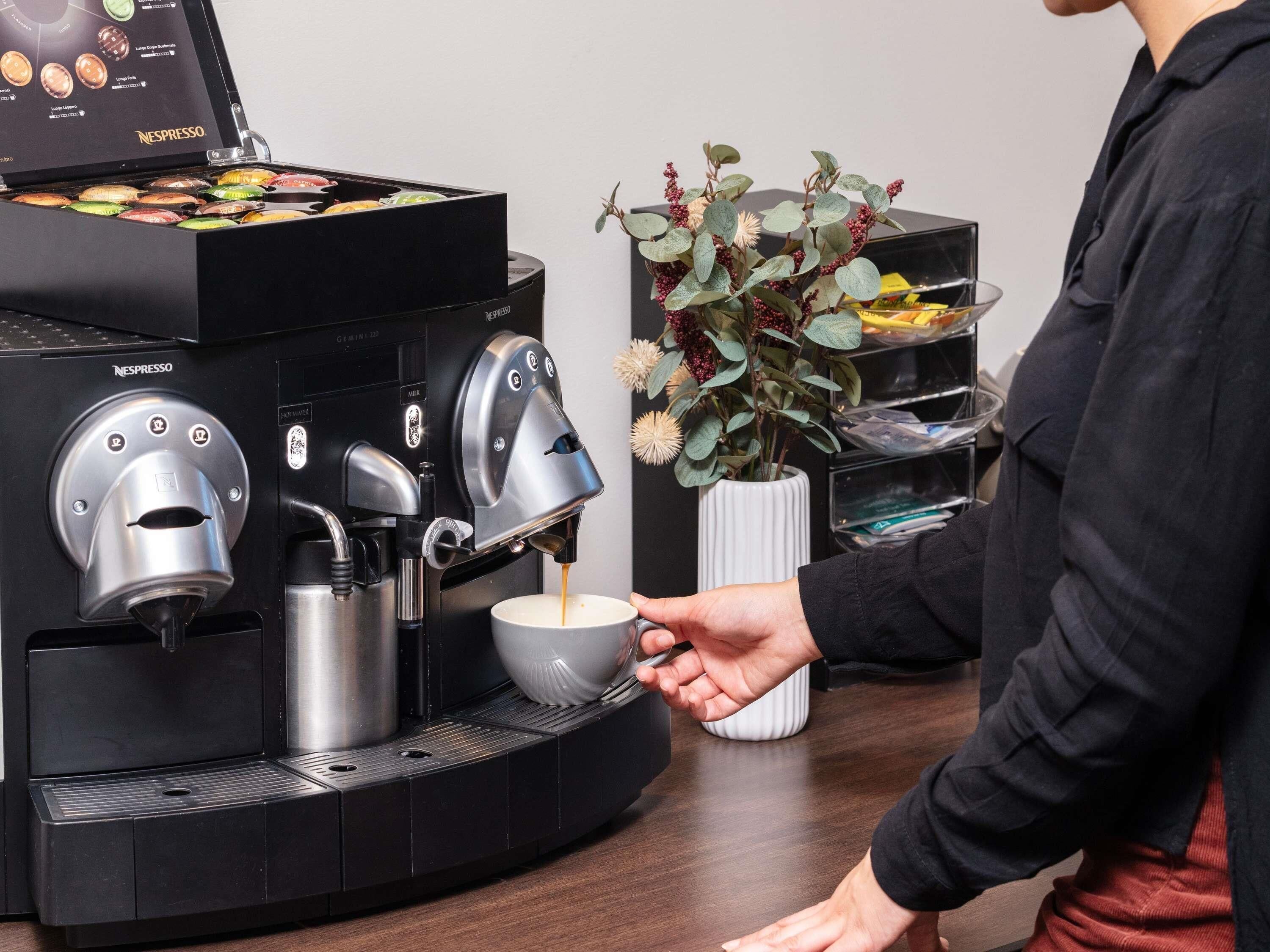 A person using a Nespresso coffee machine, with a selection of coffee pods visible and a vase with flowers on the table.