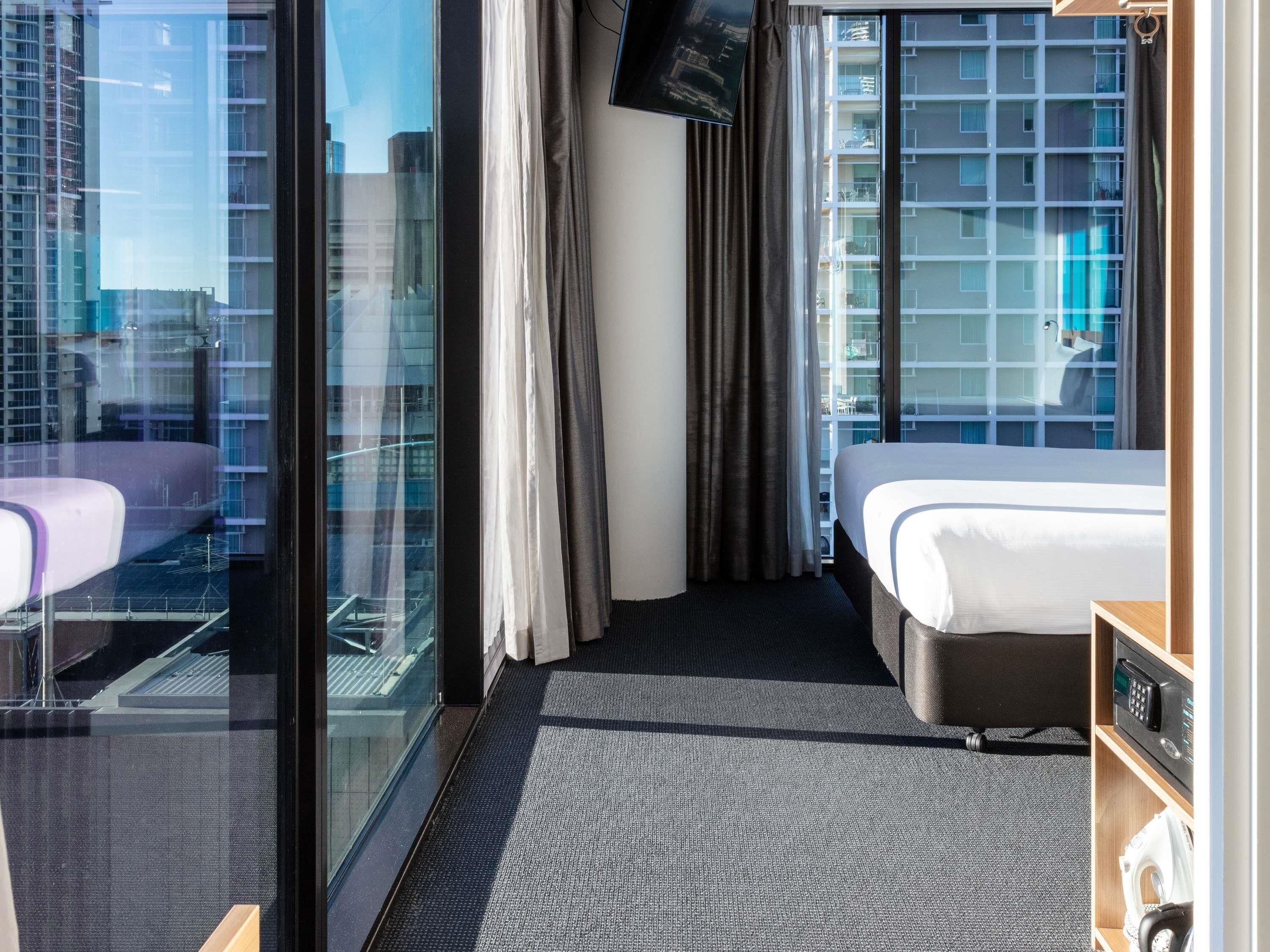 A hotel room with a bed, a desk, a TV, and a view of a building through a glass door.
