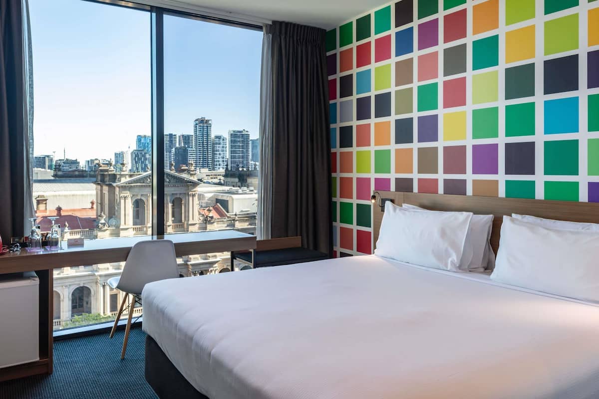 Une chambre d’hôtel avec un grand lit, un bureau, une chaise et une vue sur le paysage urbain par une grande fenêtre.