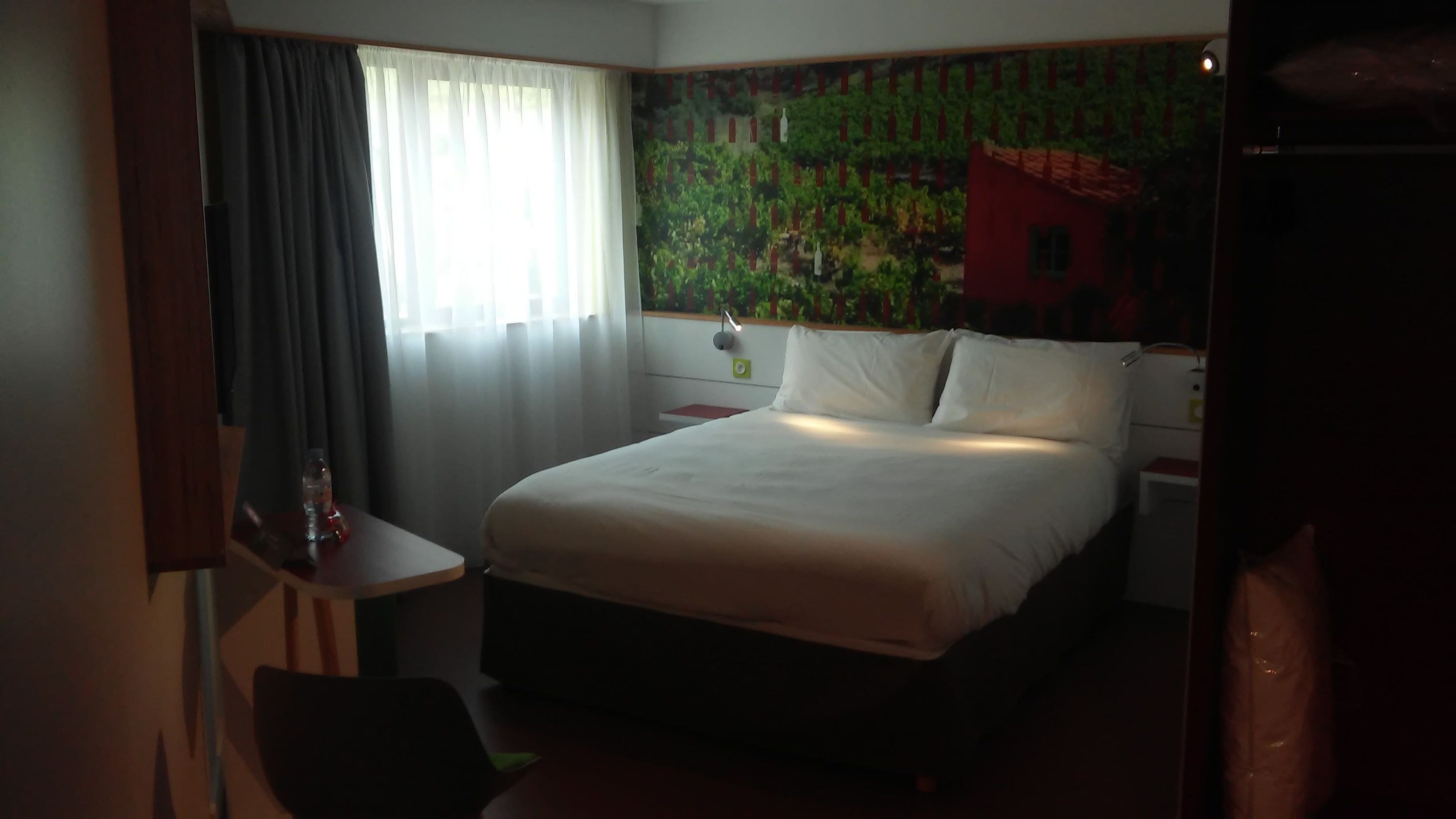 Une chambre d’hôtel comprenant un lit, un bureau, une chaise, une table de chevet et un mur orné d’une fresque représentant un vignoble.