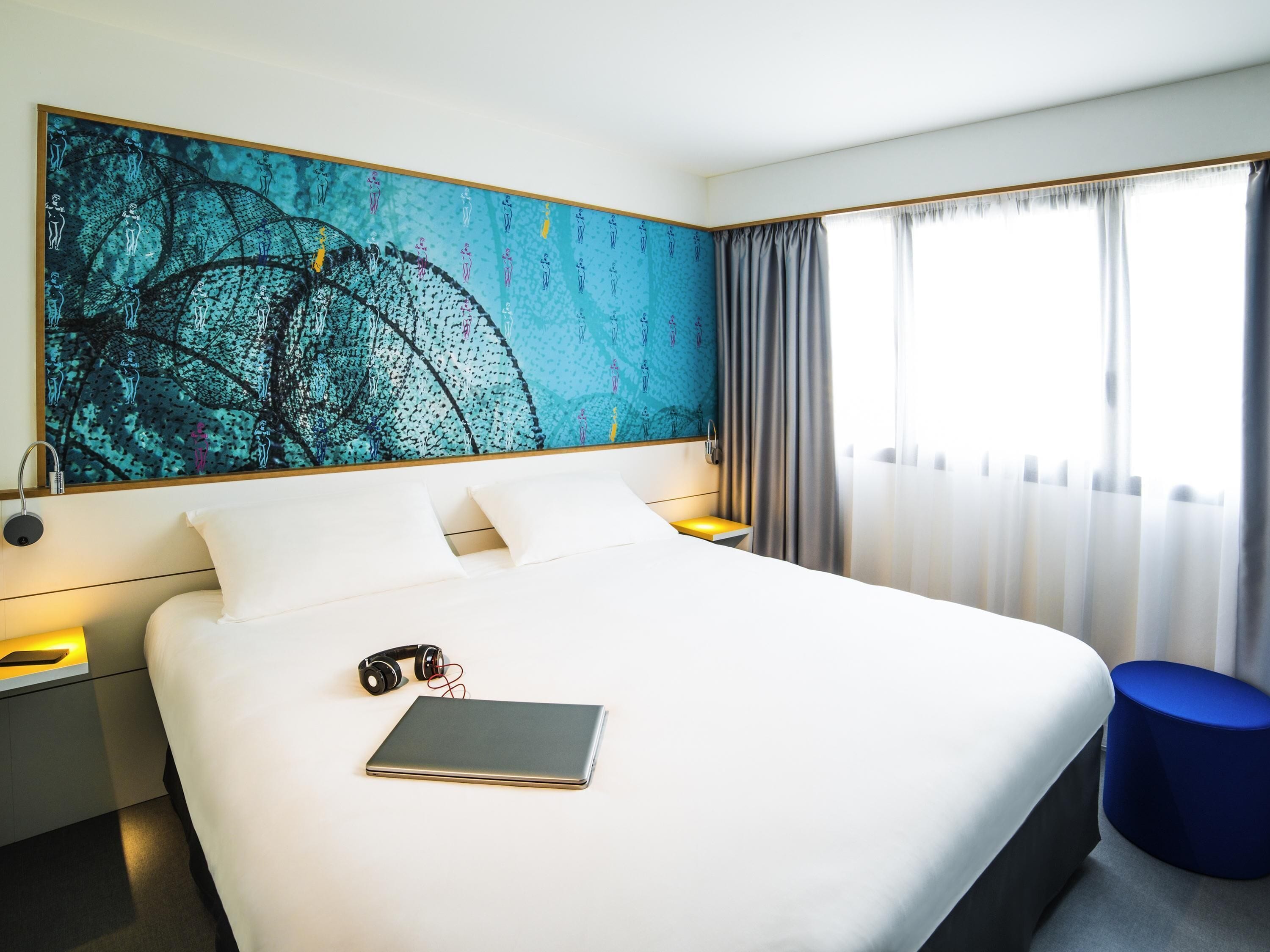 A hotel room with a large bed, a bedside table, a nightstand, a wall art piece, a window with curtains, and a blue ottoman.