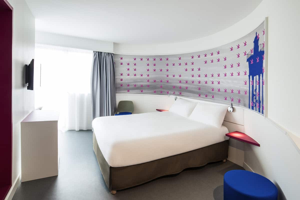 A modern hotel room with a curved bed, a desk, a chair, and a large window with curtains.